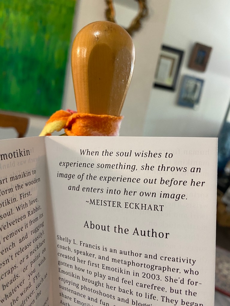 The Emotikin holds up the card deck booklet open to the page reading “When the soul wishes to experience something, she throws an image of the experience out before her and enters into her own image.” 
—MEISTER ECKHART
