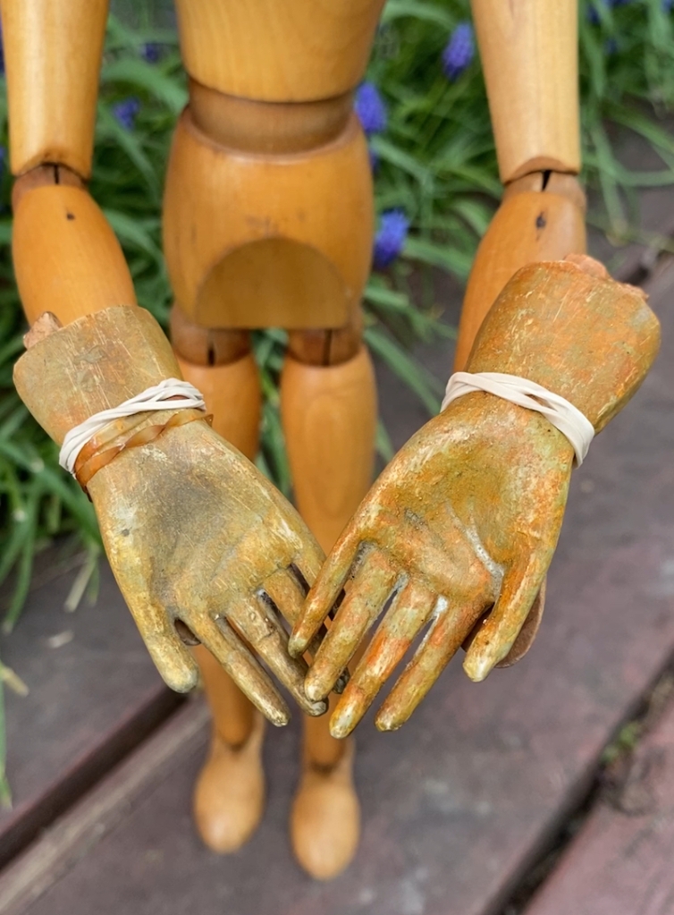 Emotikin (an art manikin) holds two wooden doll hands rubberbanded on the wrists. Green and blue grape hyacinths are in the background. 